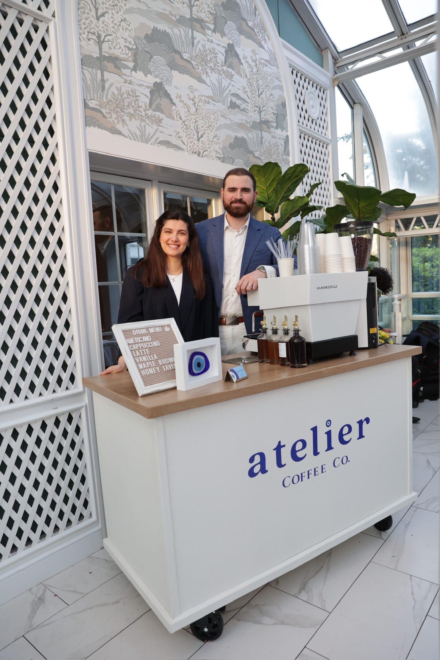 A man and a woman smiling behind the Atelier Coffee Co. cart in a glass conservatory, with the drinks menu and the nazar on the counter
