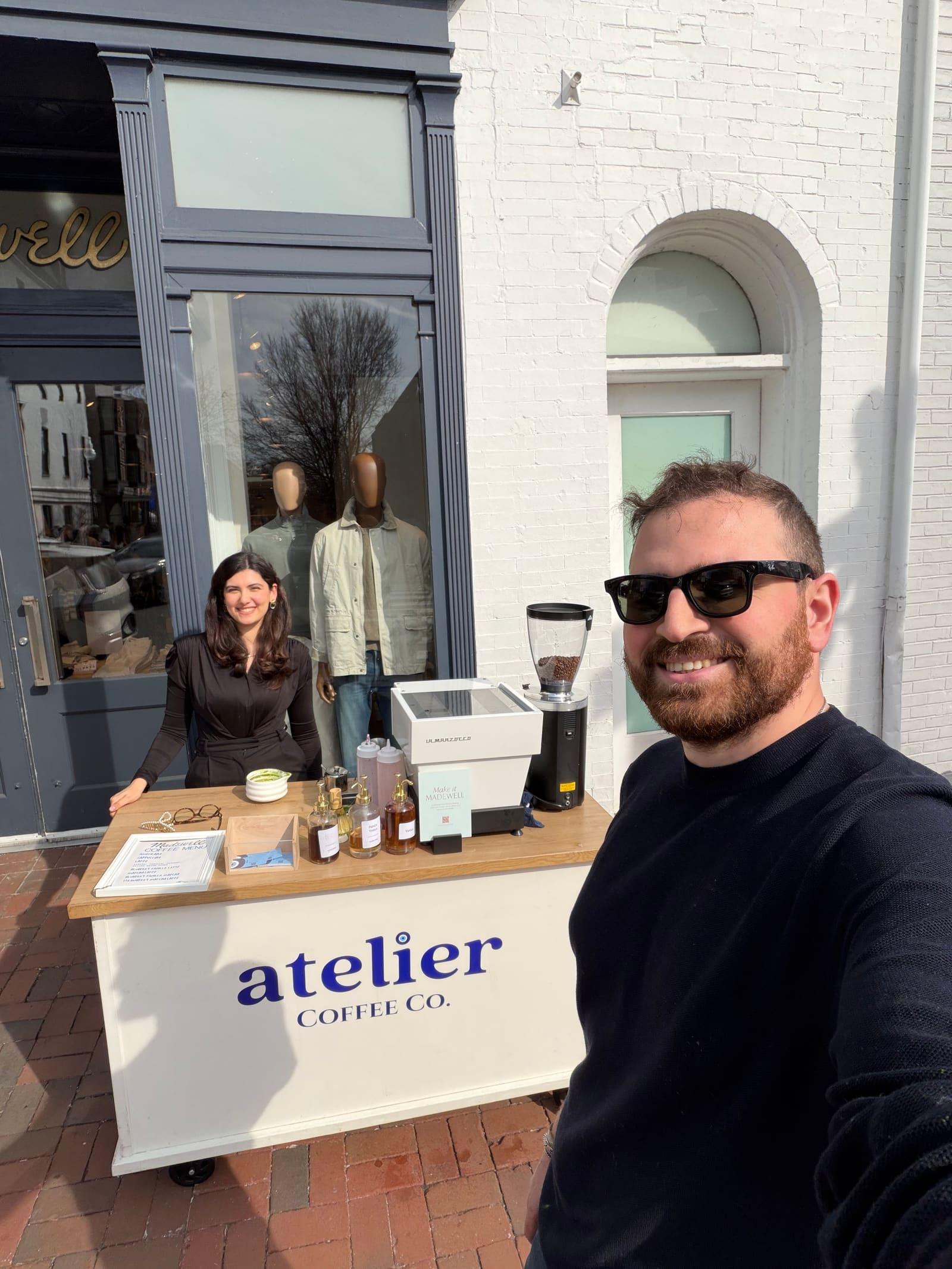 A selfie of a man in sunglasses outside a Madewell storefront, with a woman smiling behind the Atelier Coffee Co. cart