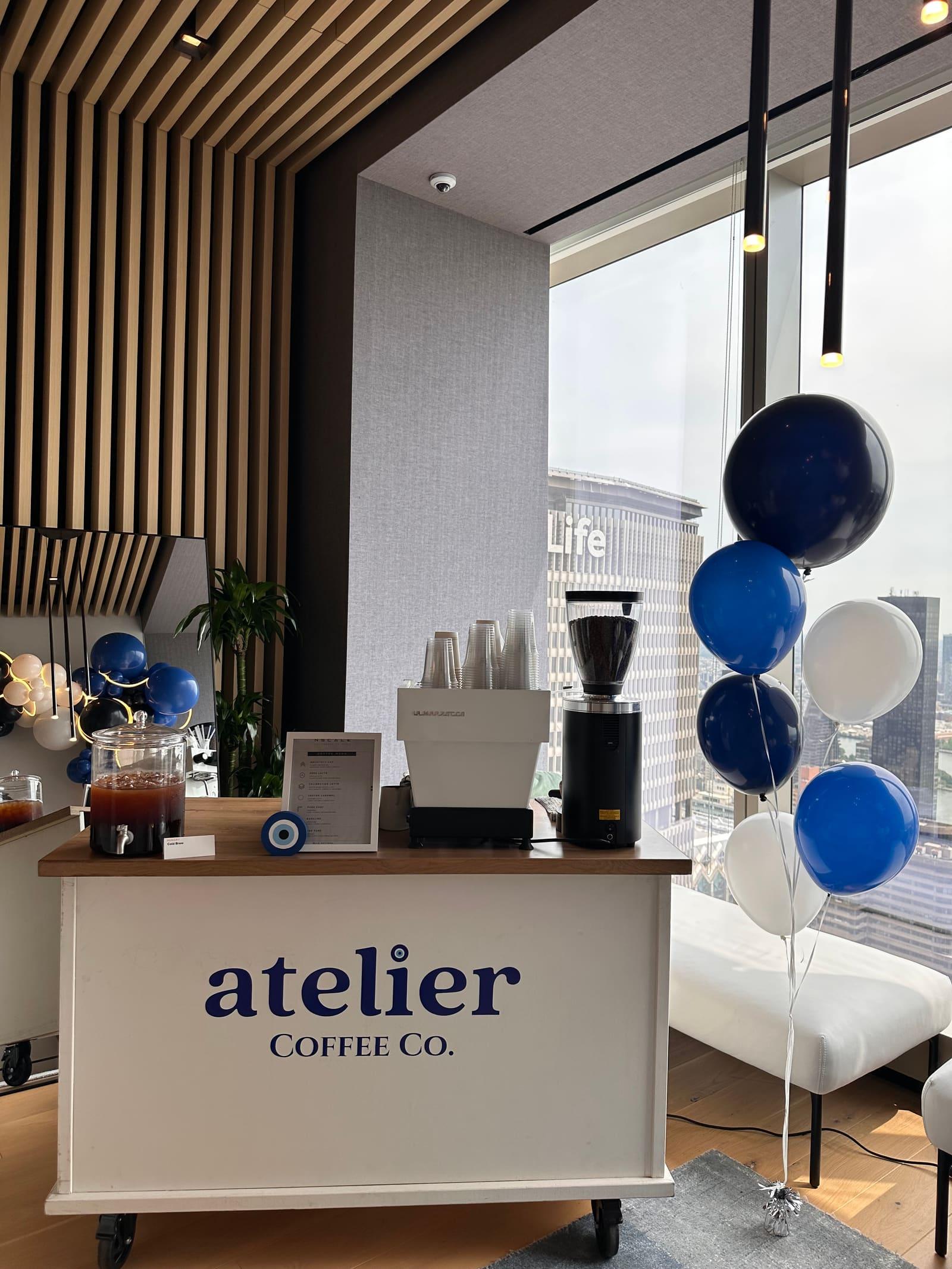 The Atelier Coffee Co. cart beside bunches of blue and white balloons, with city towers through the window
