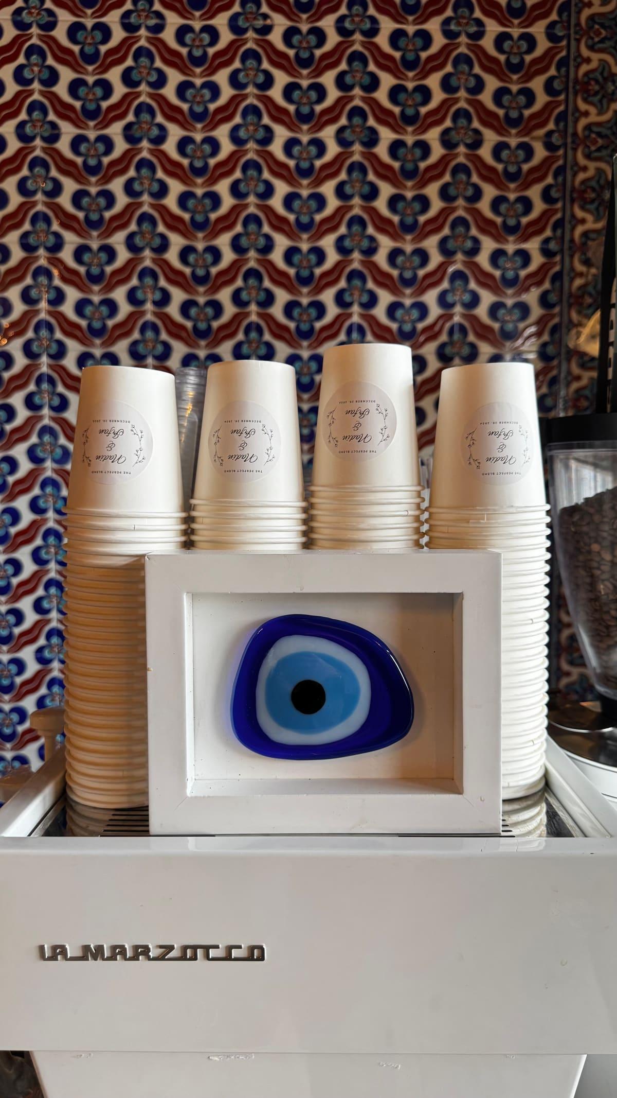 A stack of white cups and the blue nazar on an espresso machine, in front of a patterned Turkish textile