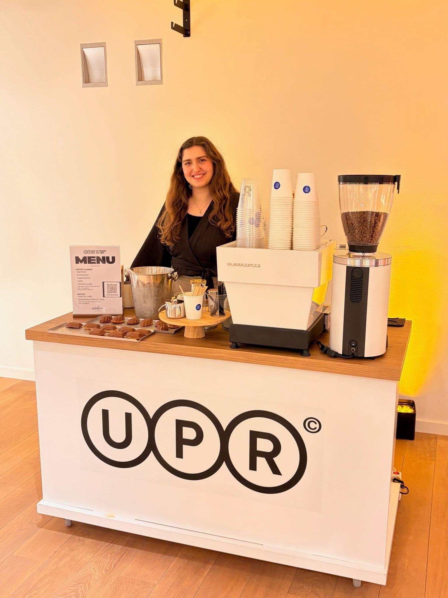 An Atelier barista behind a branded espresso cart in a warmly lit room