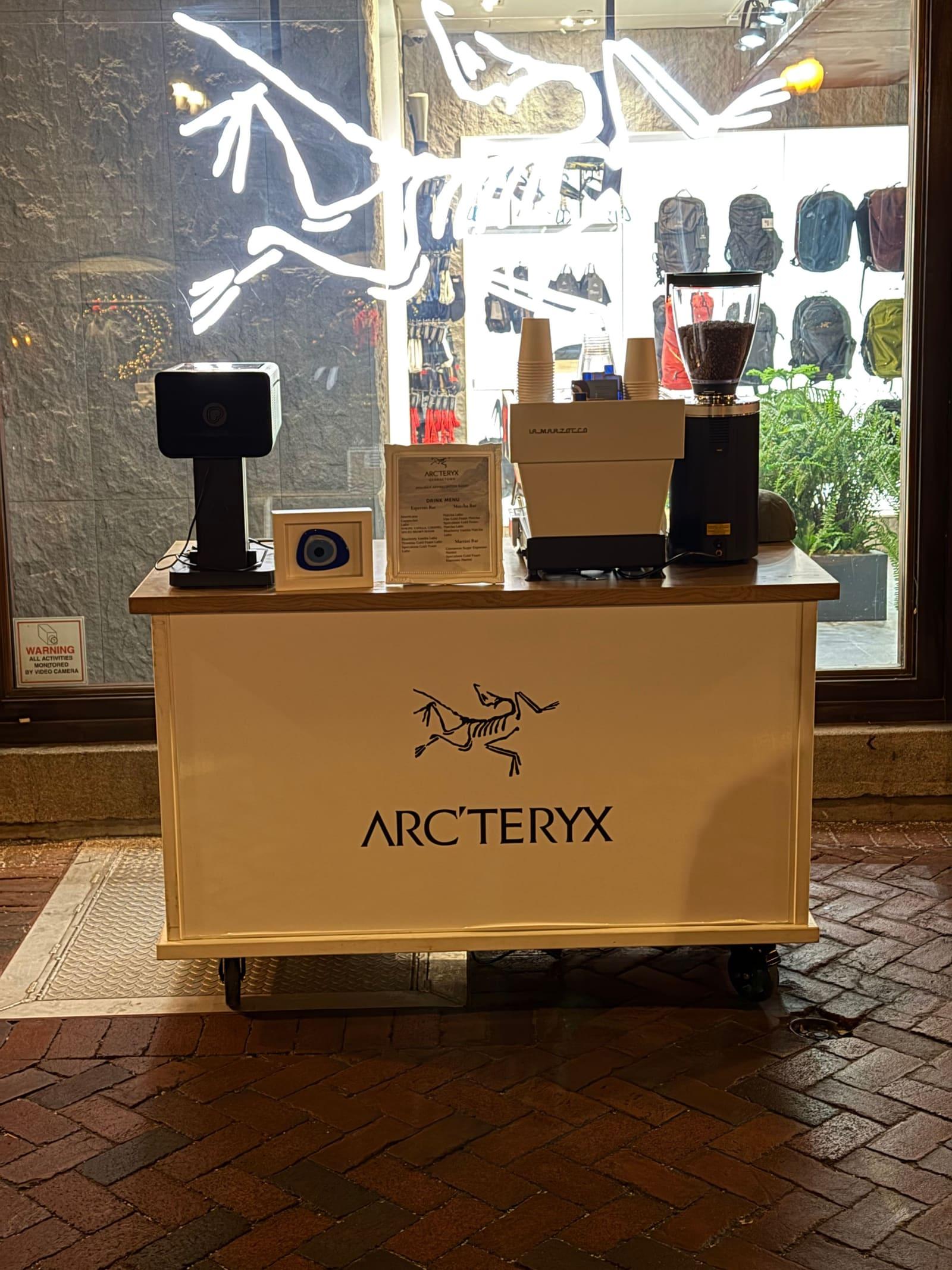 An espresso cart wrapped in tan Arc’teryx branding beside a shop window lit with the Arc’teryx bird