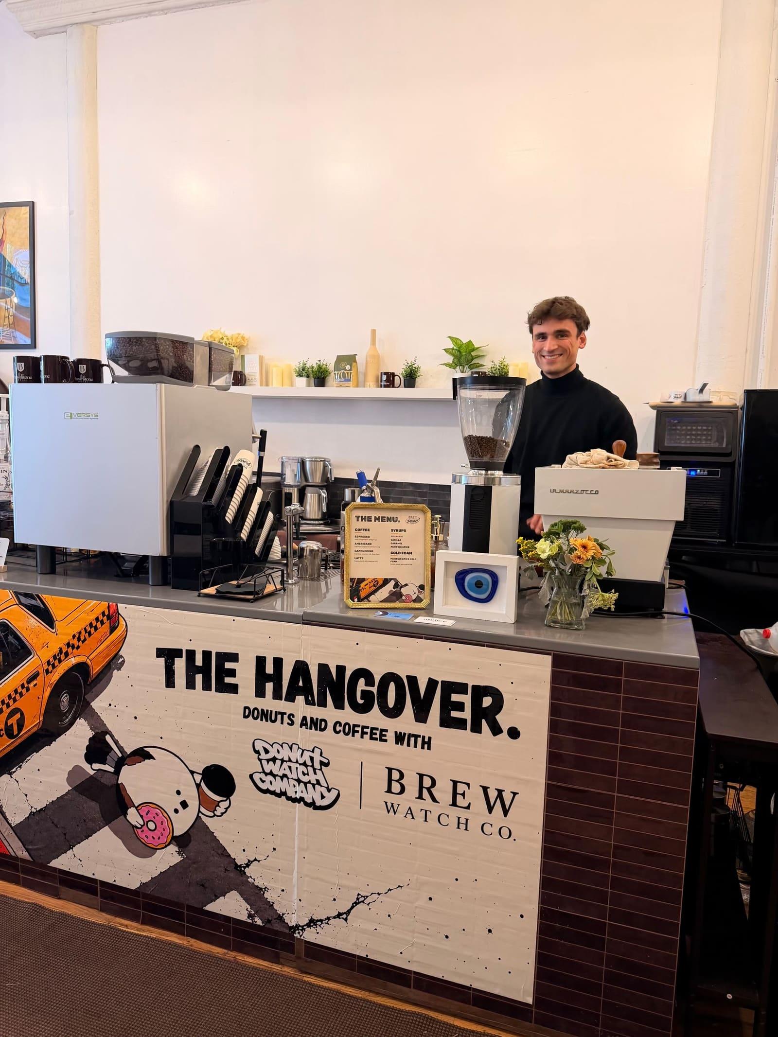 An Atelier barista behind a bar wrapped in custom artwork, serving donuts and coffee