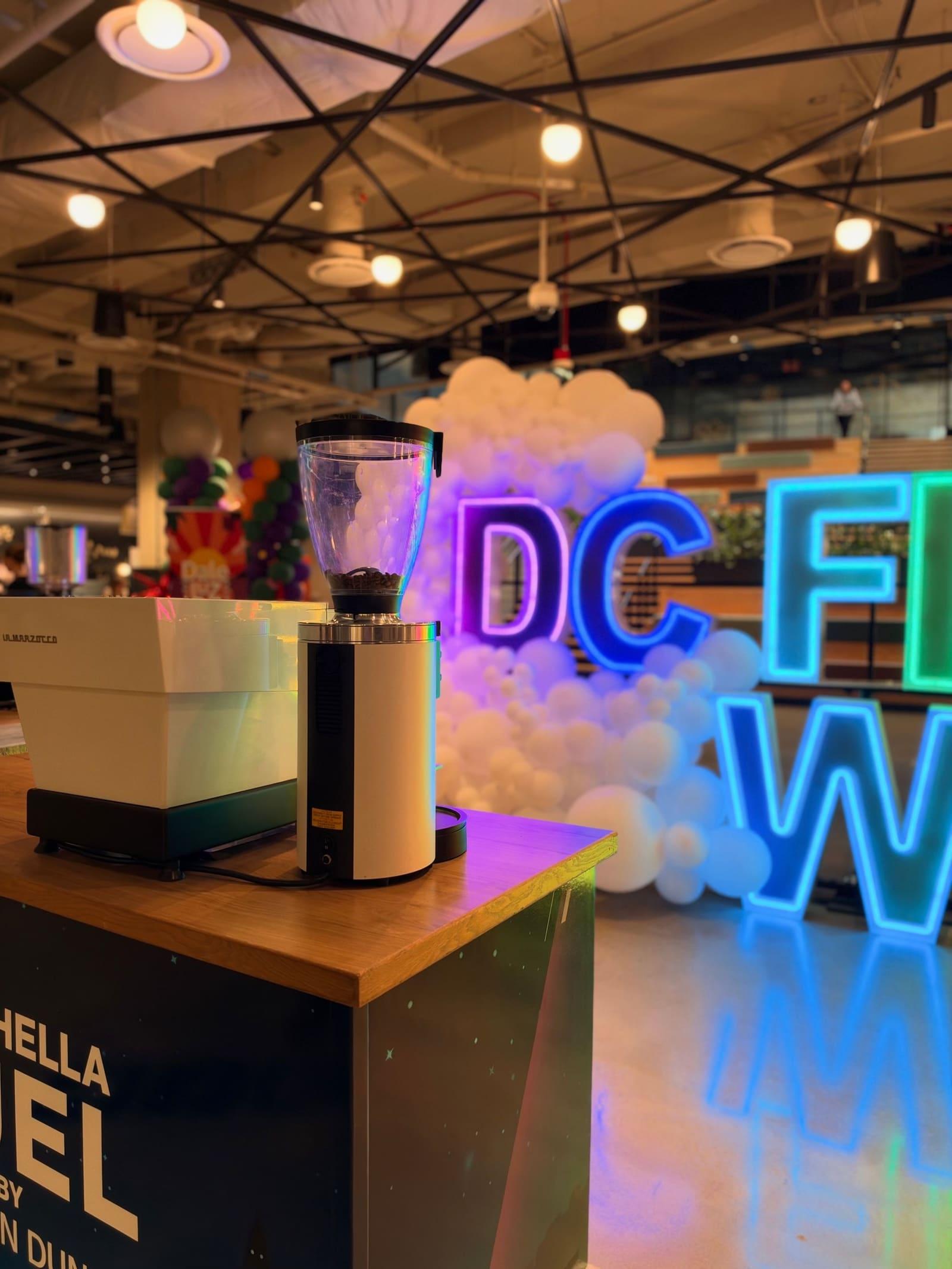 A branded espresso cart in front of a glowing neon conference sign and balloons