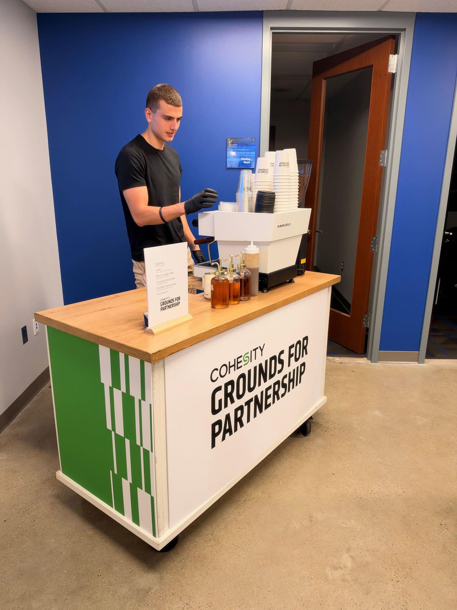 An Atelier barista at an espresso cart wrapped as “Grounds for Partnership”, in an office with a blue wall