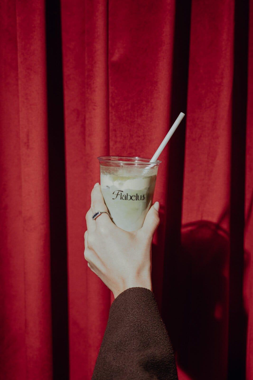An iced matcha latte in a logo‑printed cup, held up against a red velvet curtain