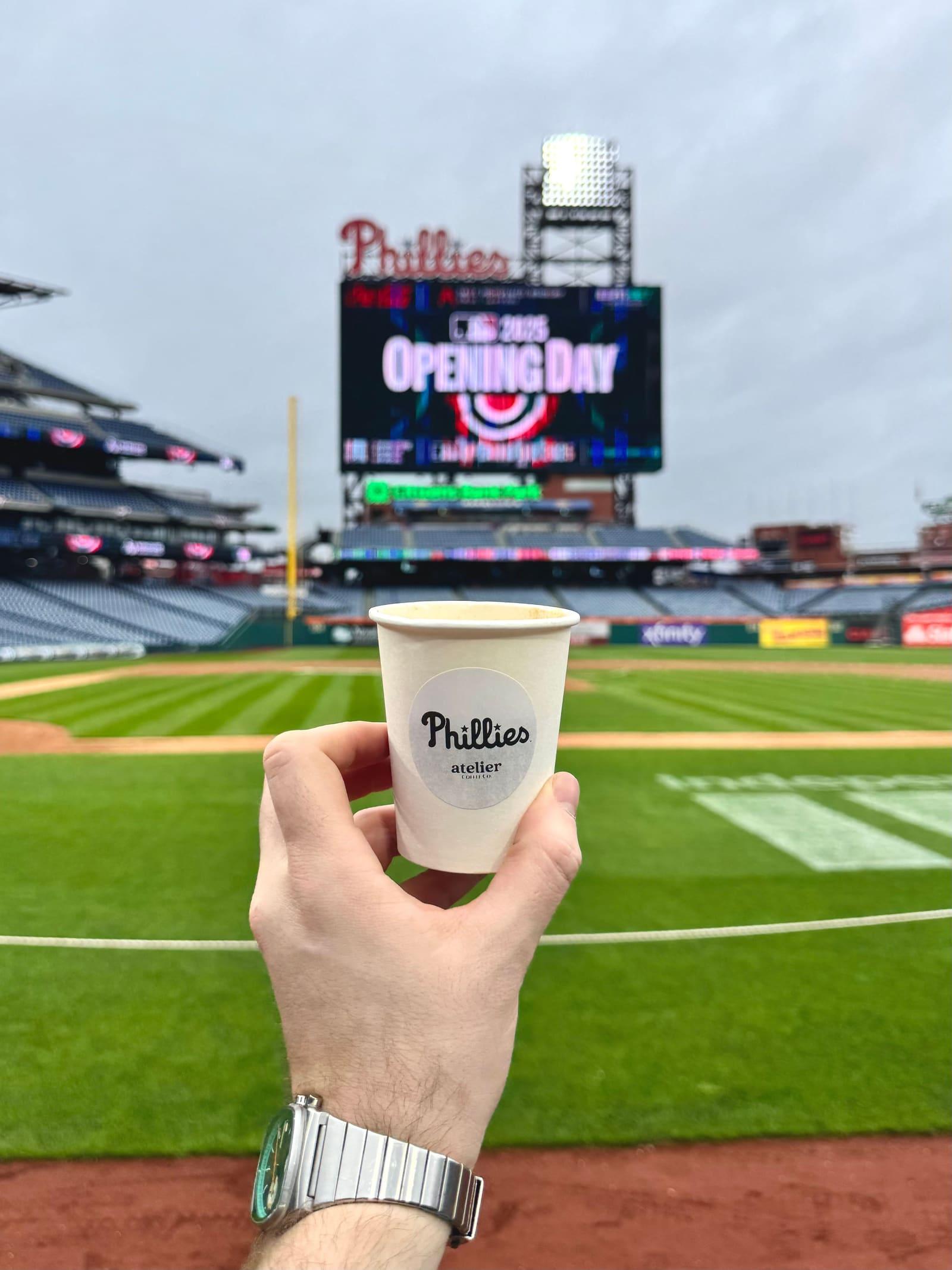 A coffee in a Phillies cup held up over the field, with the Opening Day scoreboard behind