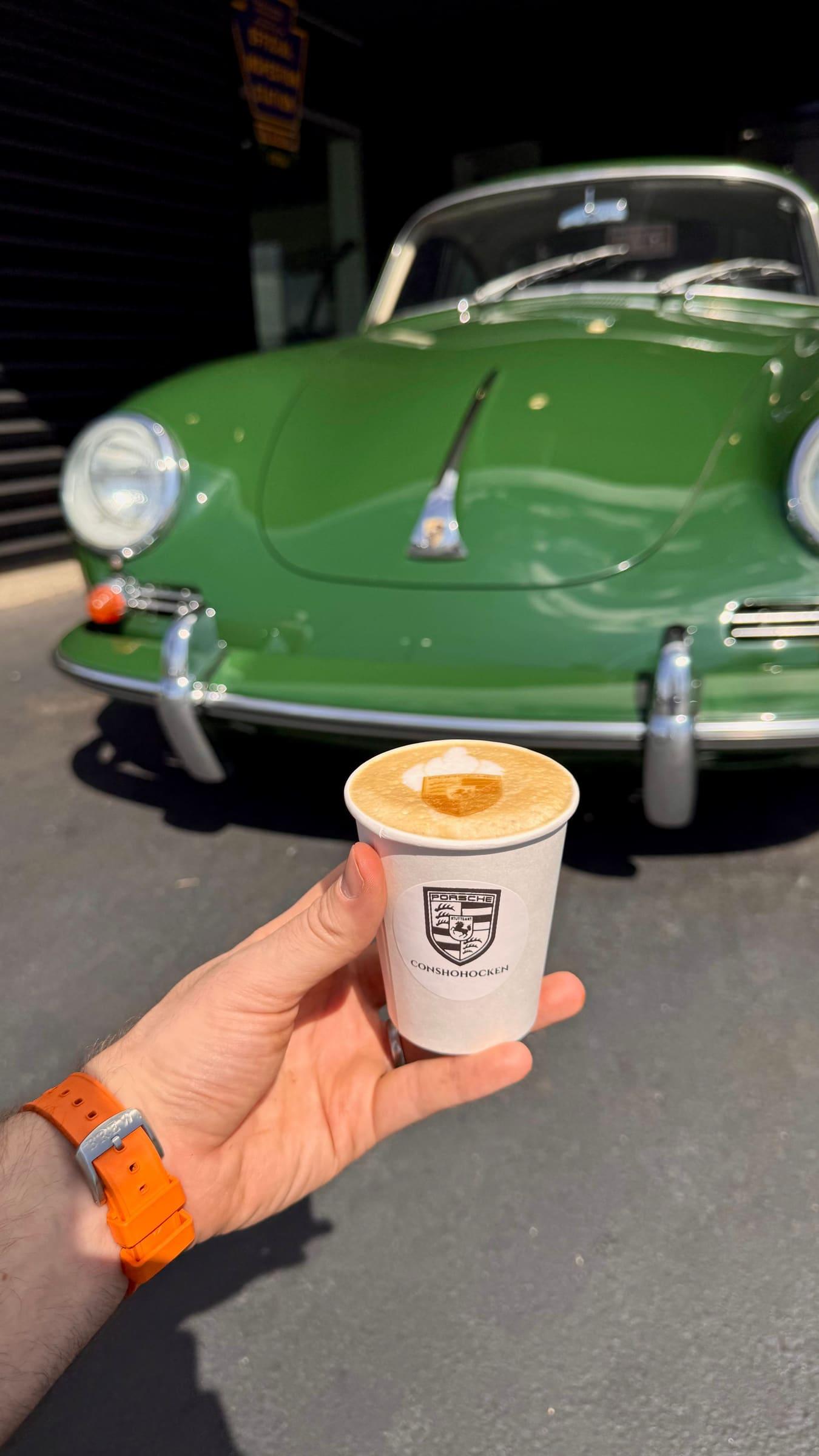 A coffee in a cup with the Porsche crest, held up in front of a green vintage Porsche 356