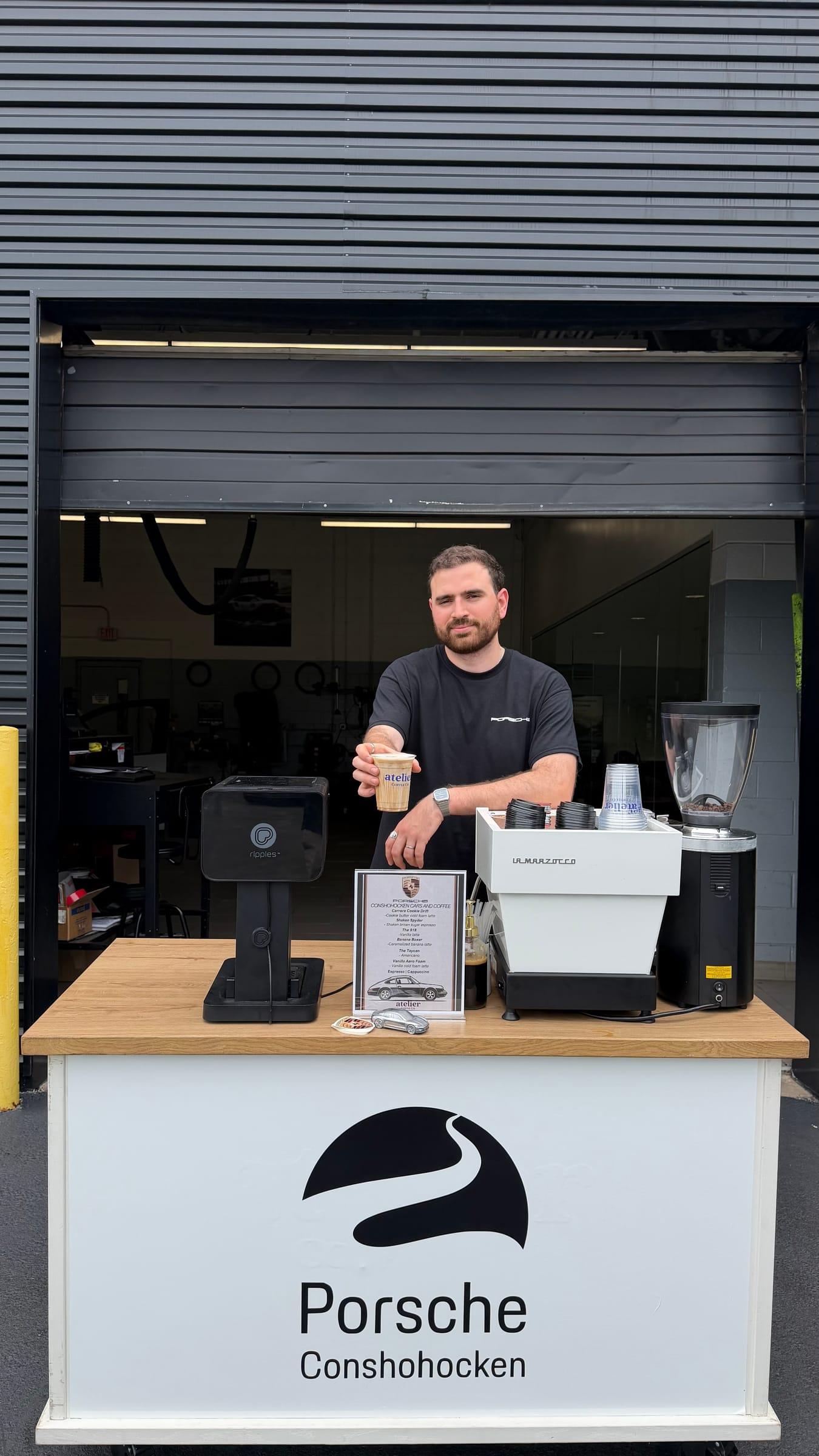An Atelier barista holding a fresh drink behind a Porsche Conshohocken branded espresso cart