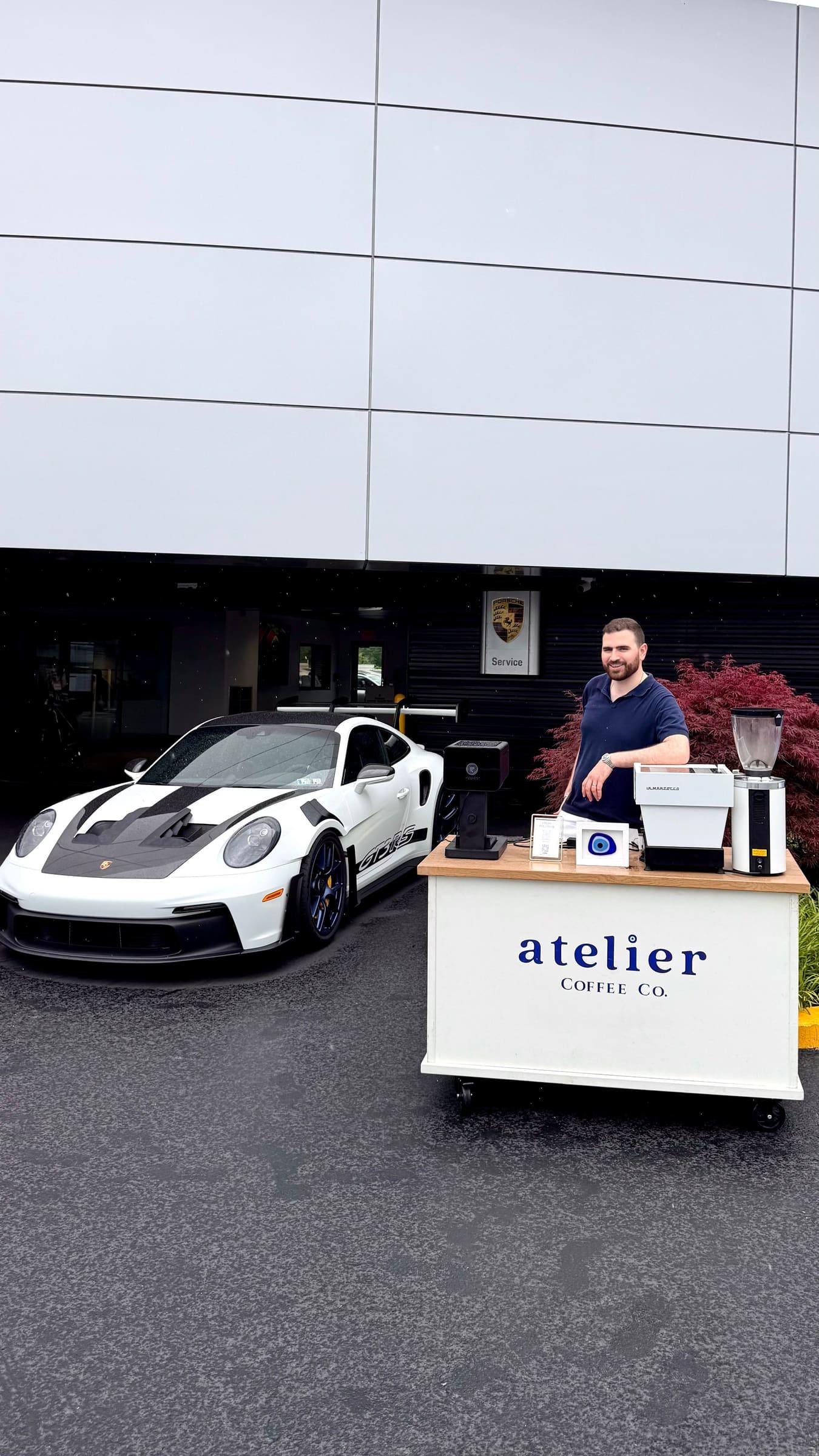 The Atelier cart beside a white Porsche GT3 RS outside the dealership, with a barista behind it