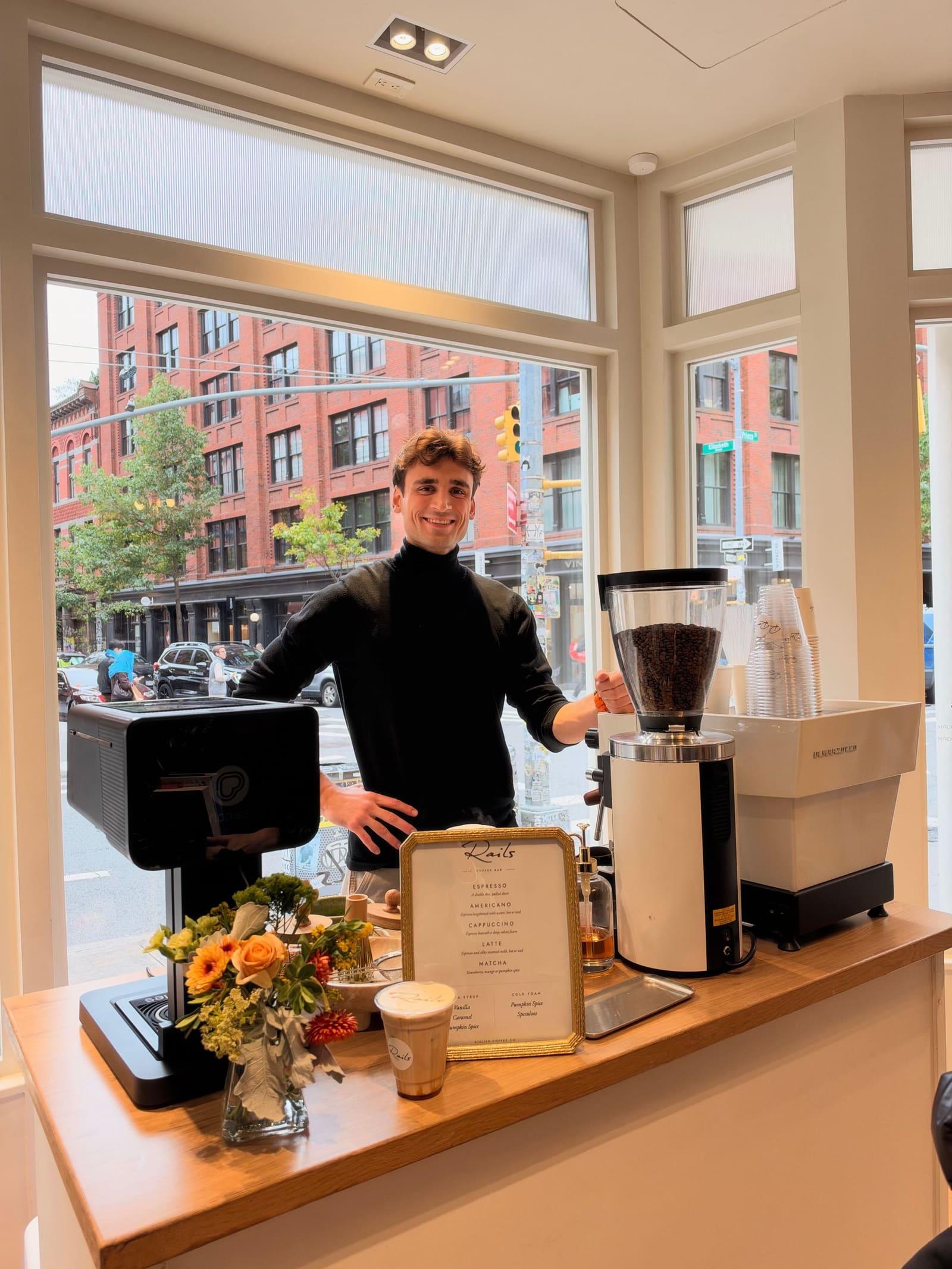 An Atelier barista at an espresso cart inside a clothing store, with brick buildings through the window behind