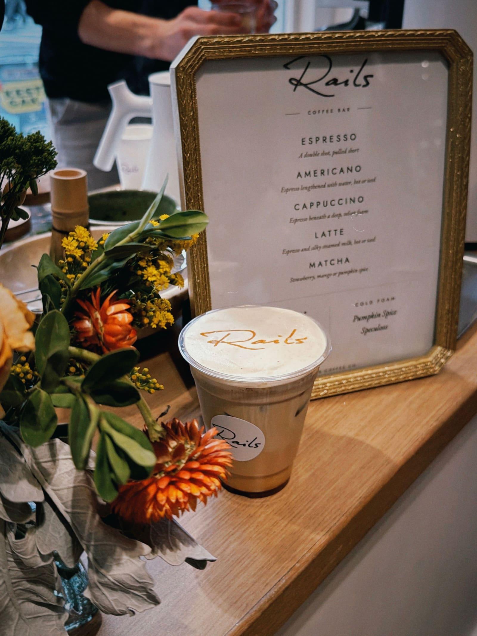 A latte in a branded cup beside a framed coffee menu and flowers