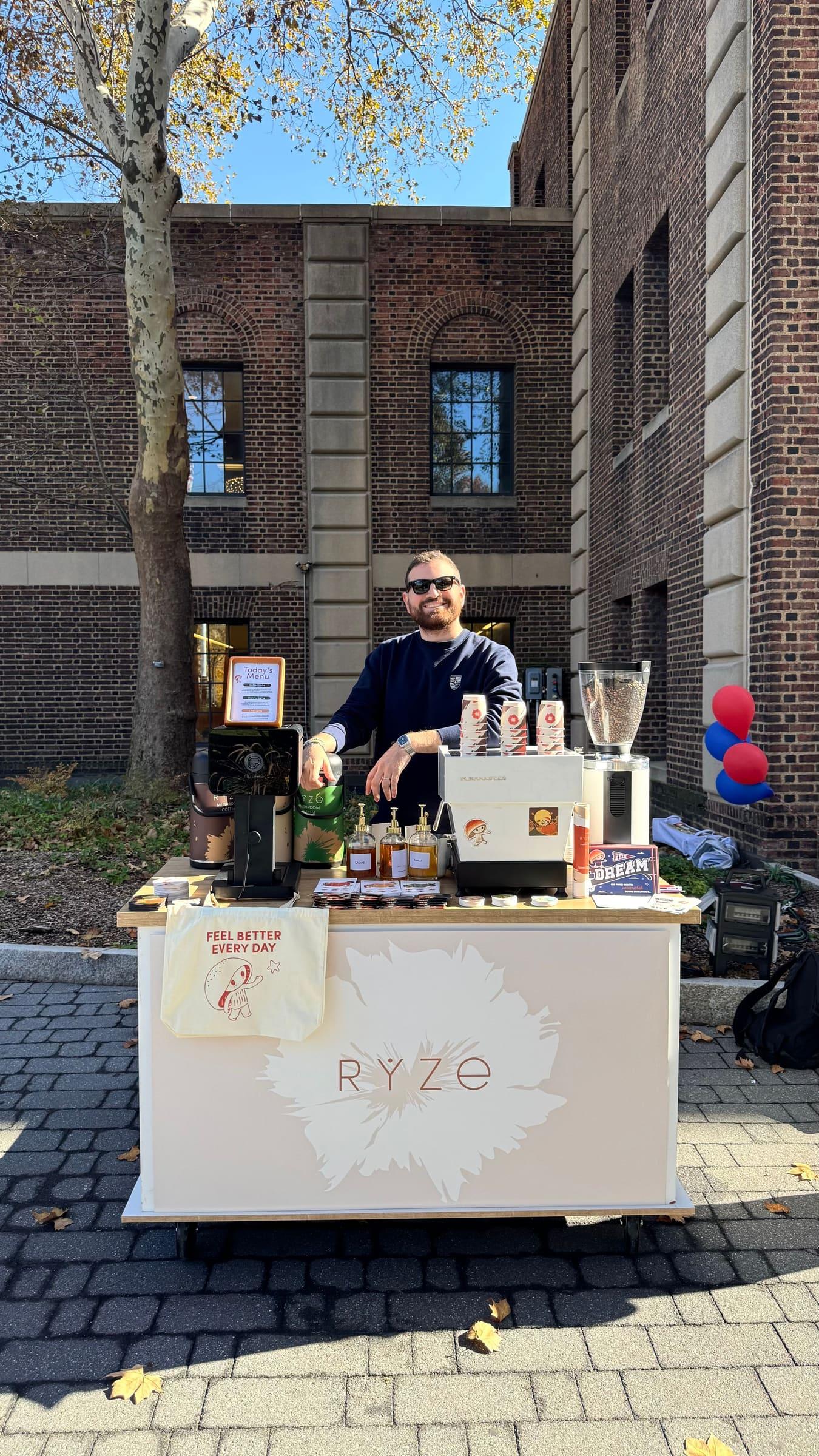 An Atelier barista behind a branded cart outdoors on a brick plaza in the fall